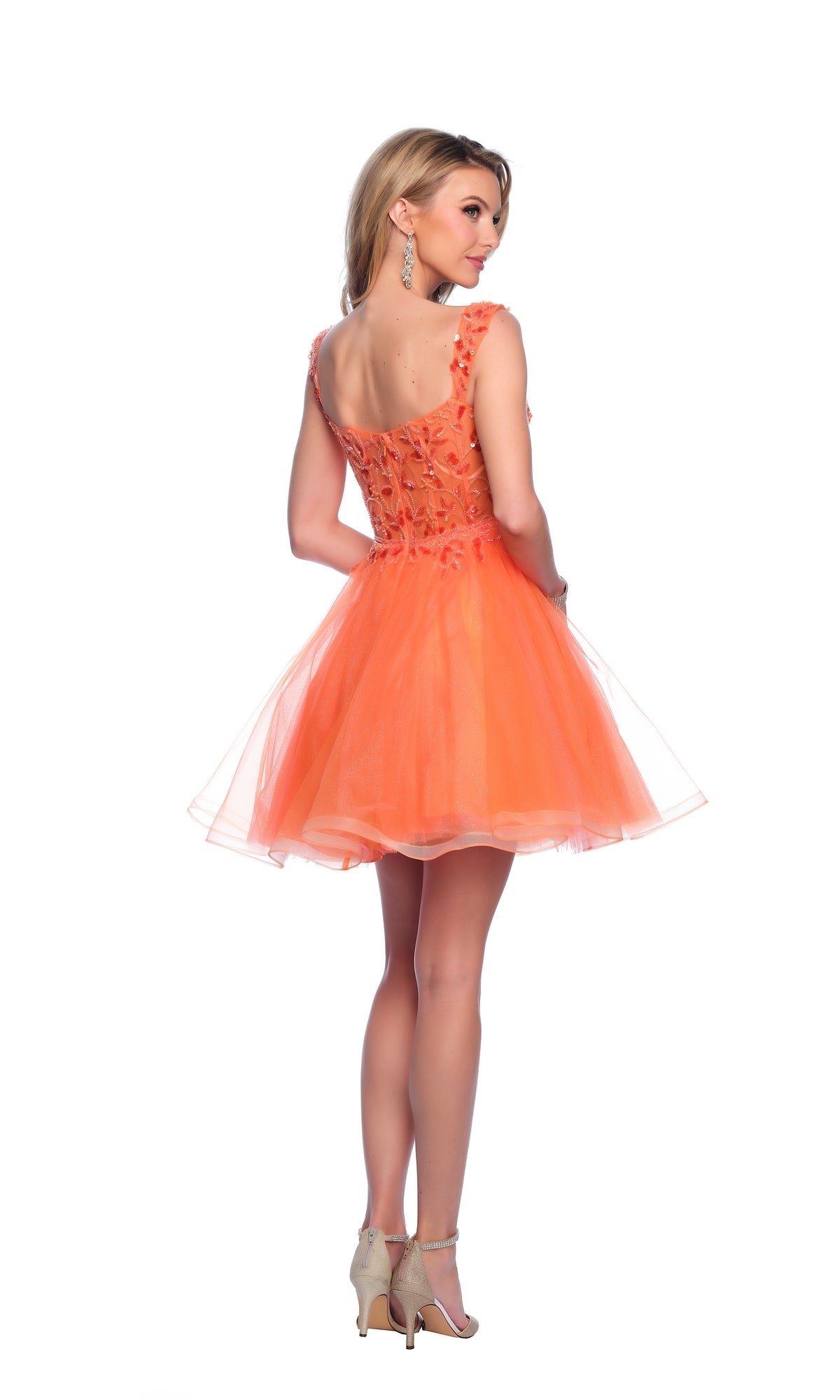 A woman models the Orange Homecoming Dress 12038, a short, sleeveless dress with floral embellishments and high heels, posing against a white background while looking over her shoulder.